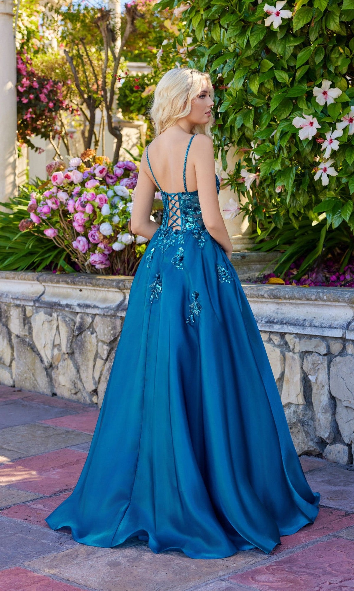 Wearing the Juliet JT2617U long prom dress in teal with floral embroidery and a lace-up back, a woman stands outdoors among blooming flowers and greenery, facing away.